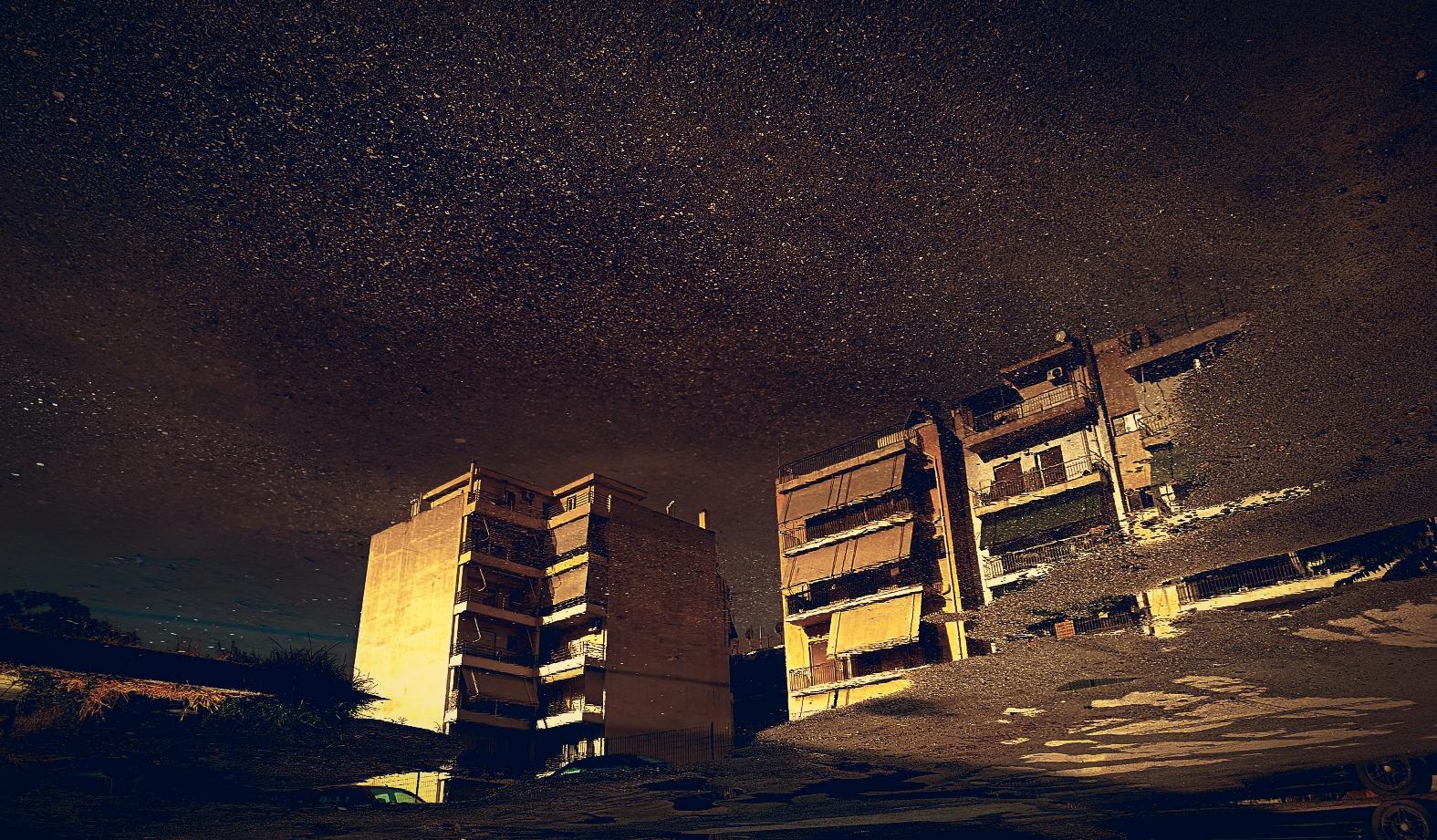 Residential buildings lit by an already hot sun, reflected in the puddling water of a sinking part of the street. The image is rotated upside-down. 
There's a hint of blue sky in the reflection, on the left, behind the buildings. The gravel around the water is trying to imitate a starry sky. There are no starry skies in Athens, I have to invent them.
The image is a low-res copy but if you are curious enough you can imagine the lives of the people in the stacked apartments, behind the awnings that fend off the sun.