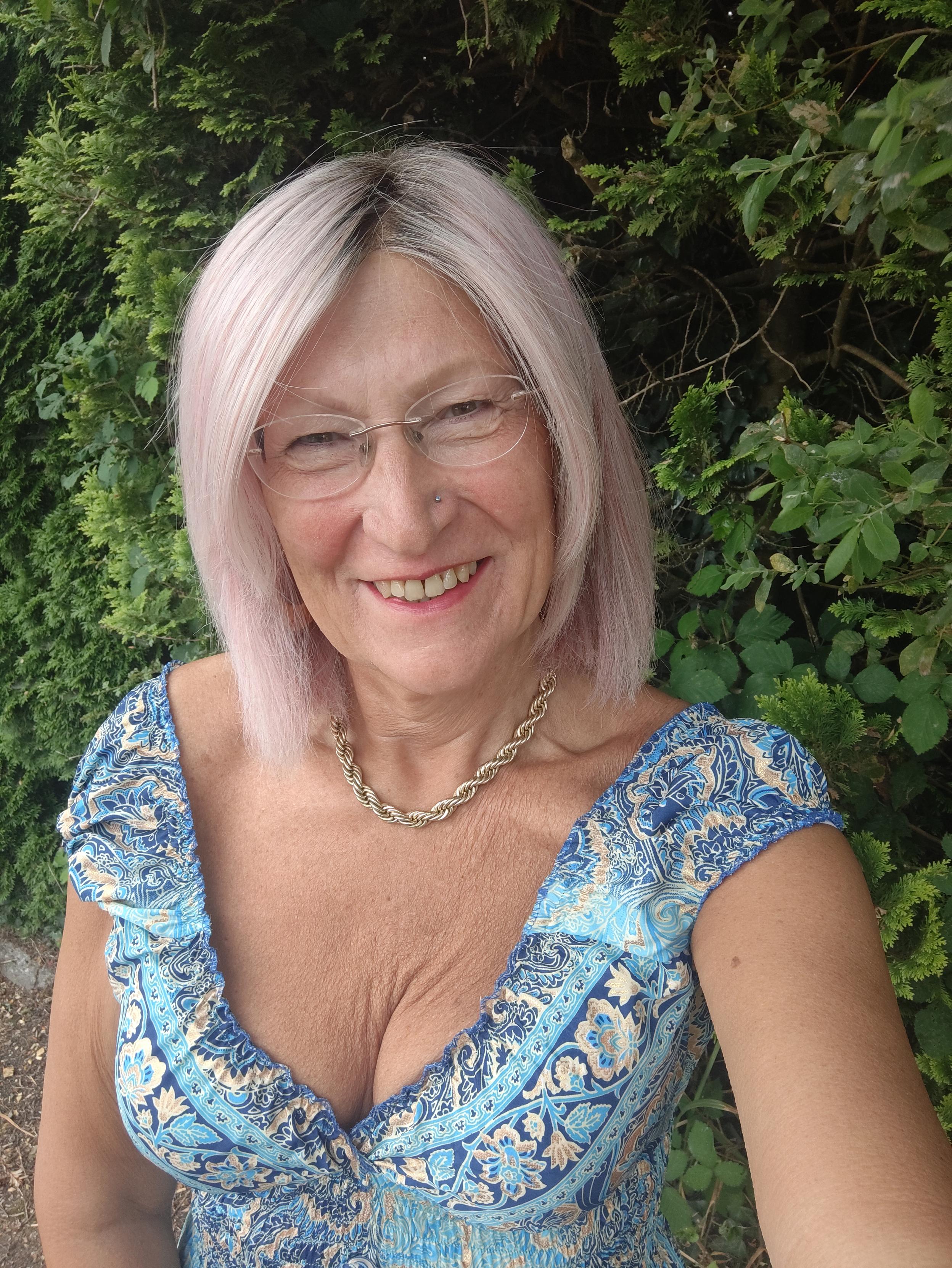 A pink haired woman standing in front of bushes wearing a blue and gold pattern low cut, sleeveless silk dress, a gold chain necklace and gold hoop earrings.
