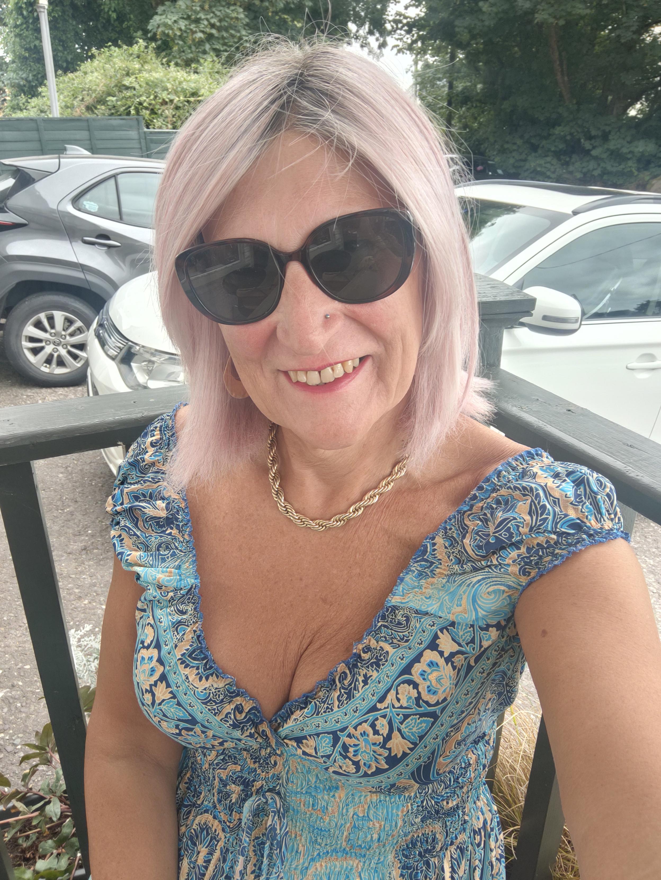 A pink haired woman sitting outside at a cafe wearing a blue and gold pattern low cut, sleeveless silk dress, a gold chain necklace, gold hoop earrings and sunglasses.
There are parked cars behind me.