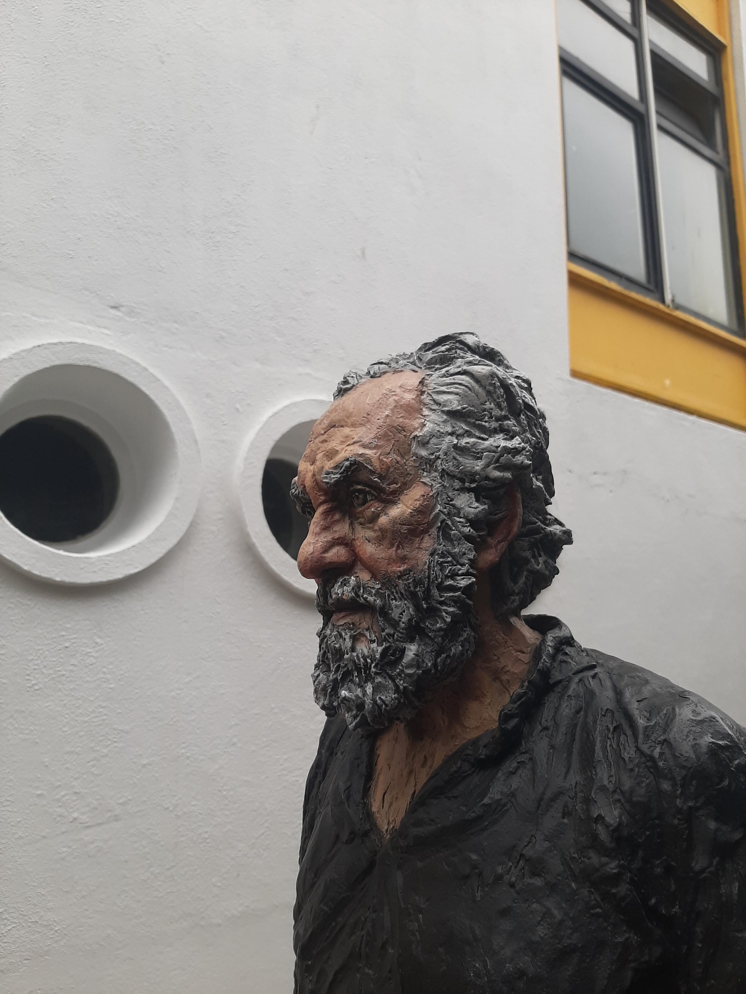 Sculpture of a grey-haired, bearded man with a troubled look on his face, seen in profile with a backdrop of the porthole windows of the festival gallery.