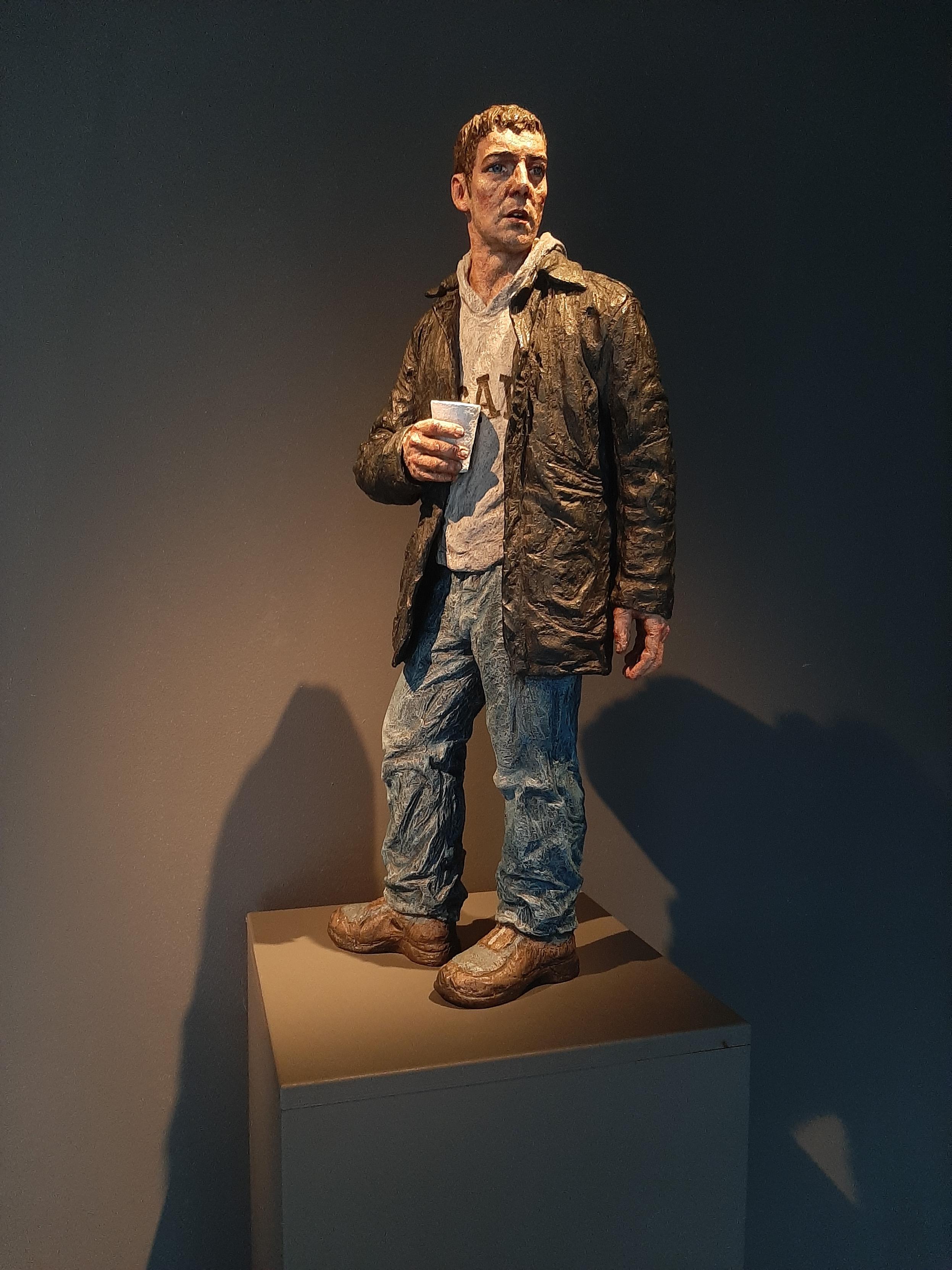 Sculpture of a young man in jeans and a 3/4 length brown leather jacket over a grey hoody, holding a plastic cup. The man is looking to one side and gives the impression of someone who has suffered hardship in his life.
