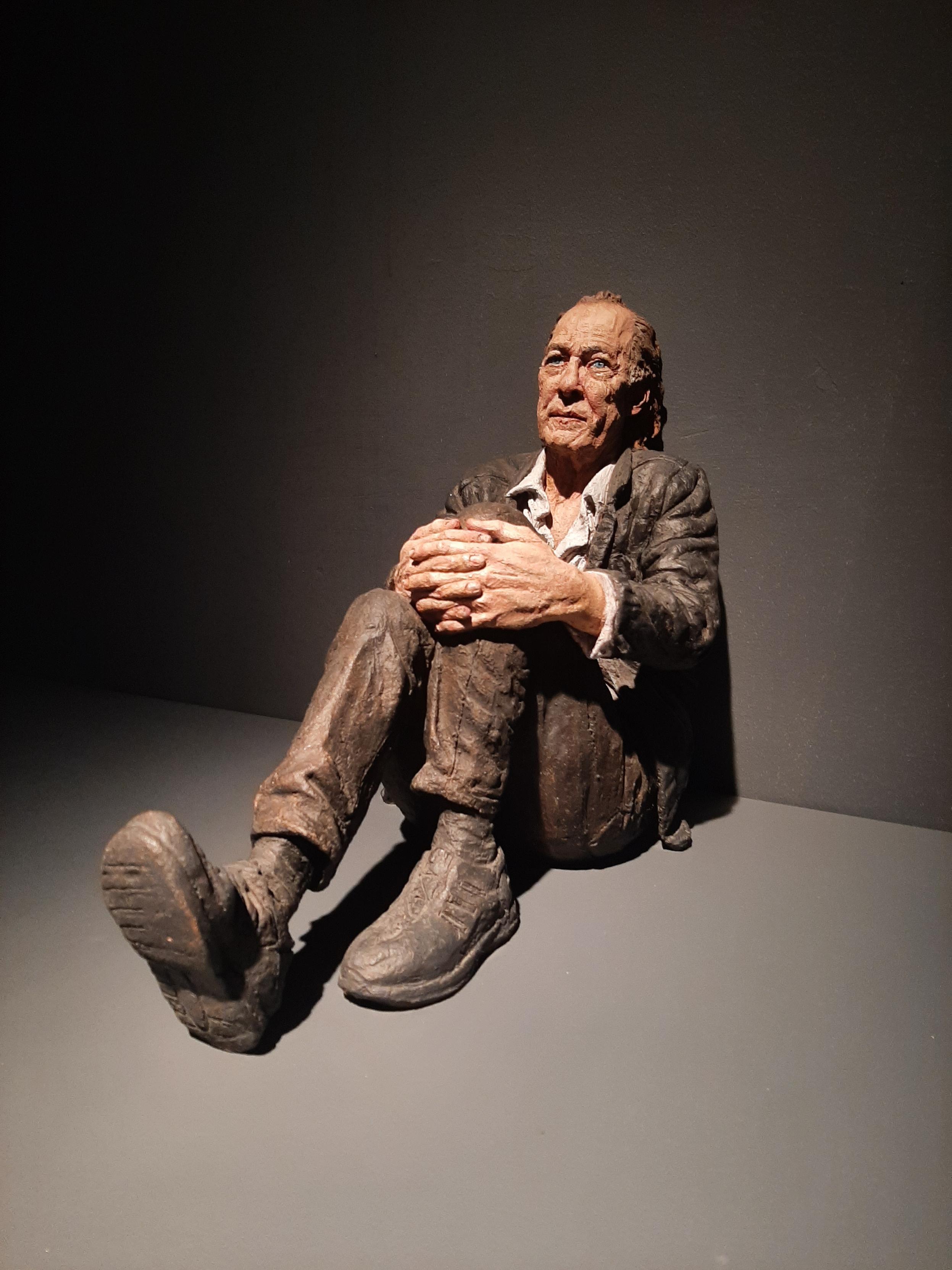 Sculpture of an older man sitting on the floor with his back against the wall, his hands entwined around one knee. The other knee is bent with the heel of the foot reading on the ground. The man looks upwards with a pensive expression like someone who has a heavy weight on his heart.