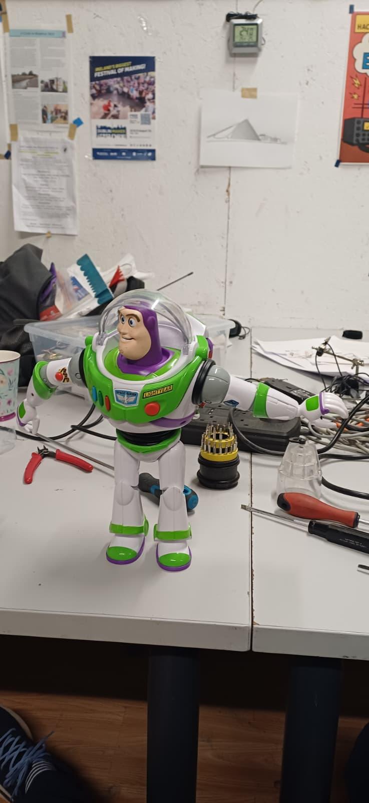 A repaired Buzz Lightyear toy standing upright on a TOG Hackerspace workbench, surrounded by screwdrivers and electronics tools.