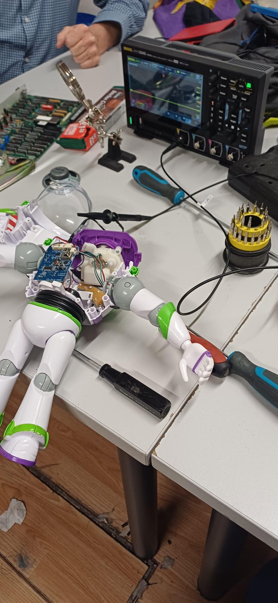 An opened Buzz Lightyear toy lying on an electronics workbench with its circuit board, internal wiring and speaker exposed beside an oscilloscope and repair tools.