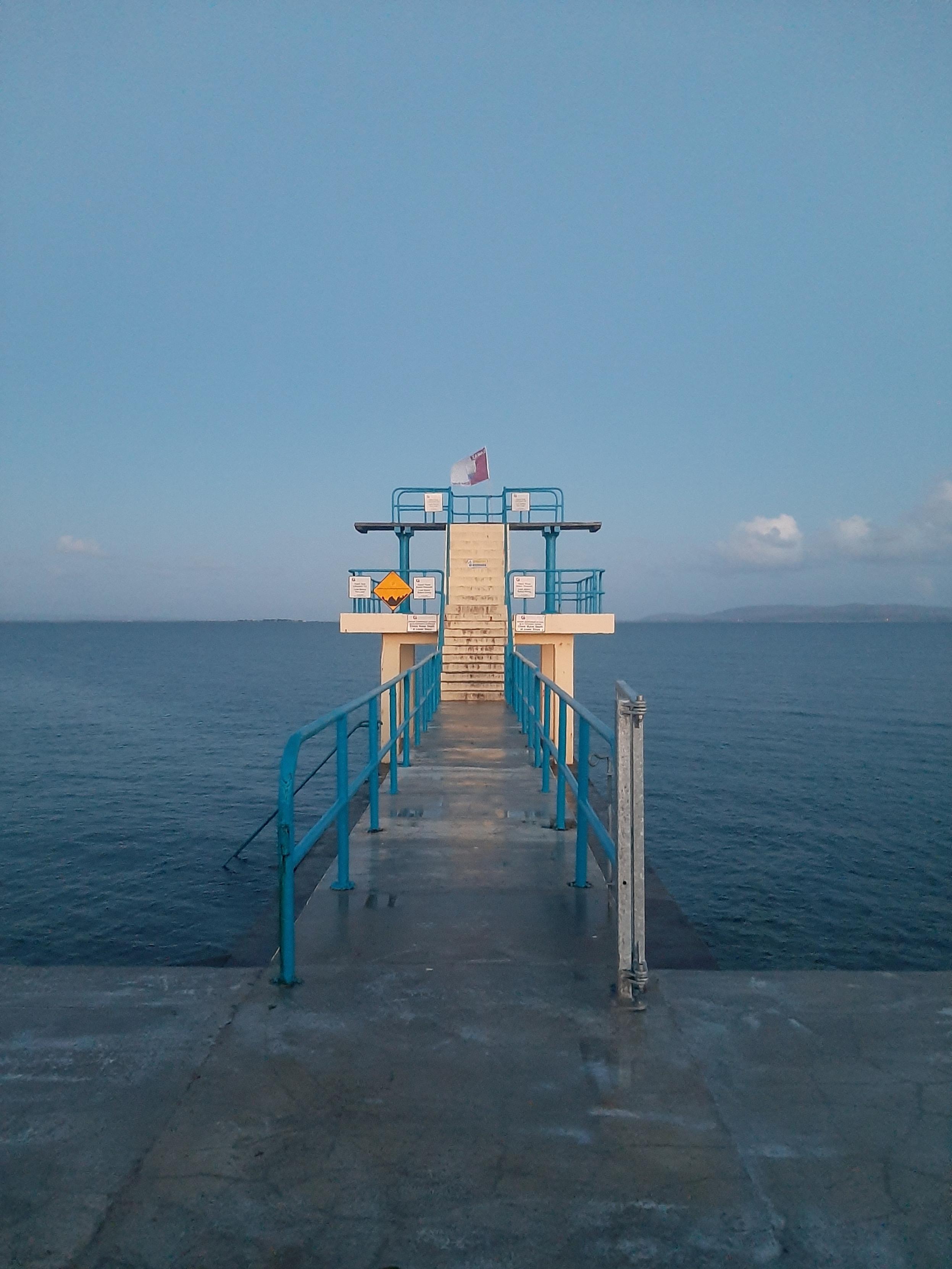 A photo of the Blackrock Diving Tower in Salthill, Galway in single point perspective looking from land towards the tower. The sky is still blue and mostly cloudless, the water is a deep blue and still, and the diving tower is yellow with blue railings. A Galway flag is flying from the top of the tower. The Clare Hills can be seen in the distance.