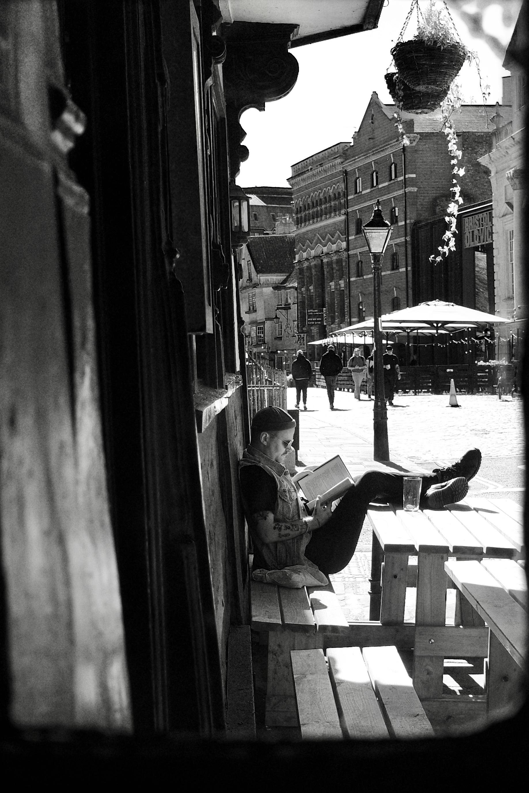 Black-and-white street photograph of a tattooed person sitting on a wooden bench outside a pub, absorbed in reading a book with their feet propped on a table. The sunlit street beyond has pedestrians, old brick buildings, a traditional lamppost, outdoor seating, and a hanging flower basket.