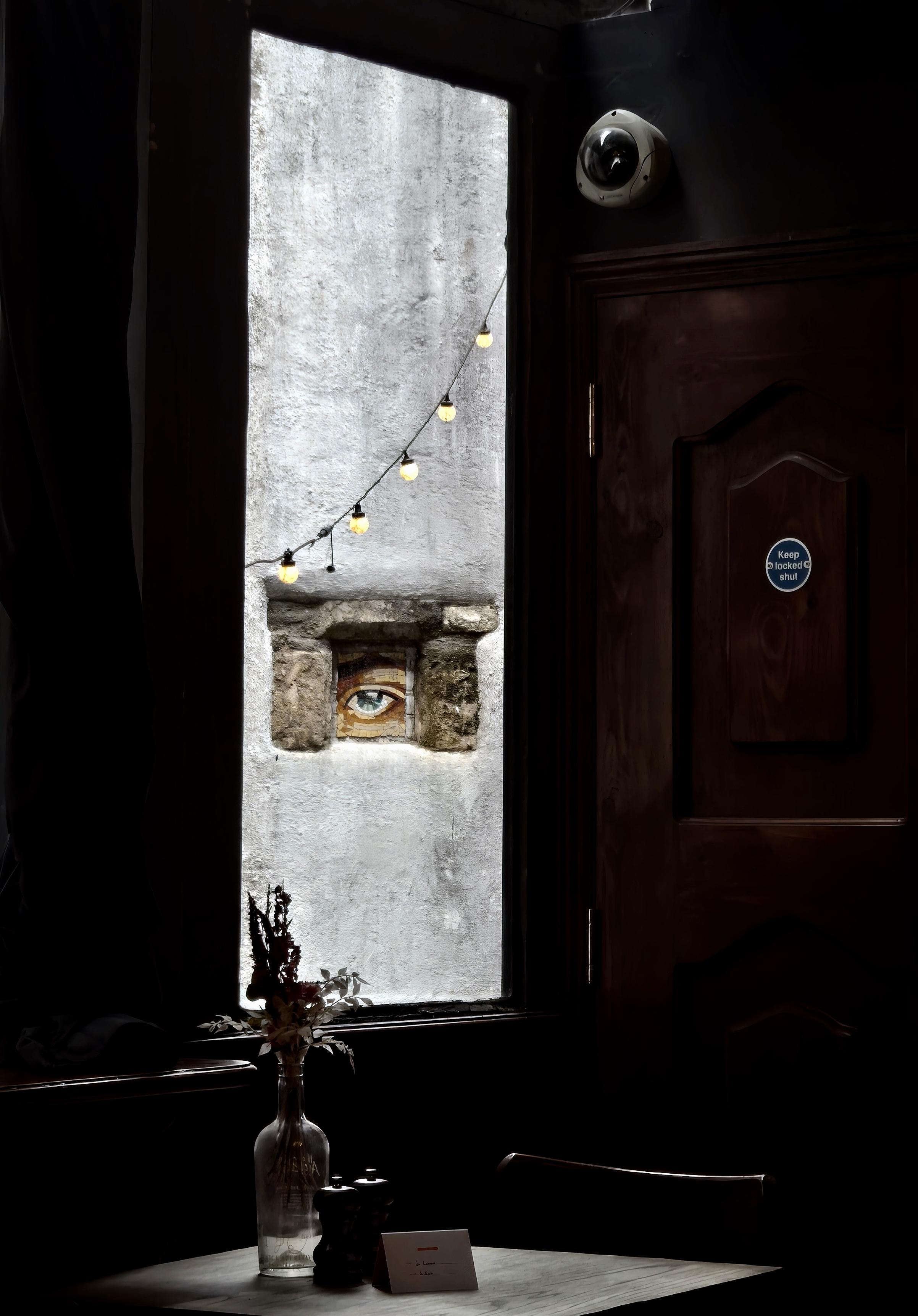 Moody interior photograph of a dark restaurant or pub. Bright daylight pours through a tall window onto a weathered exterior wall, where a small mosaic of an eye appears to peer inside. A string of glowing bulbs crosses the window, while a vase of dried flowers and condiments sit on a table in the shadowy foreground.