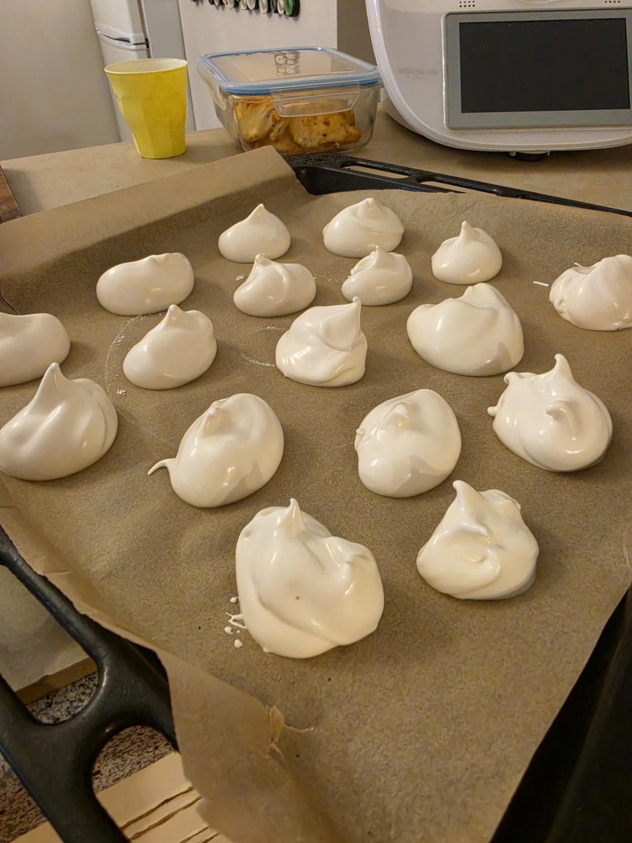 A close-up view of a baking sheet lined with parchment paper holding fresh, unbaked meringue dollops. The glossy, bright white meringue mixture forms rounded mounds with sharp, soft peaks on top. In the background on the counter sits a yellow plastic cup, a glass storage container with food inside, and a white kitchen appliance with a screen display.
