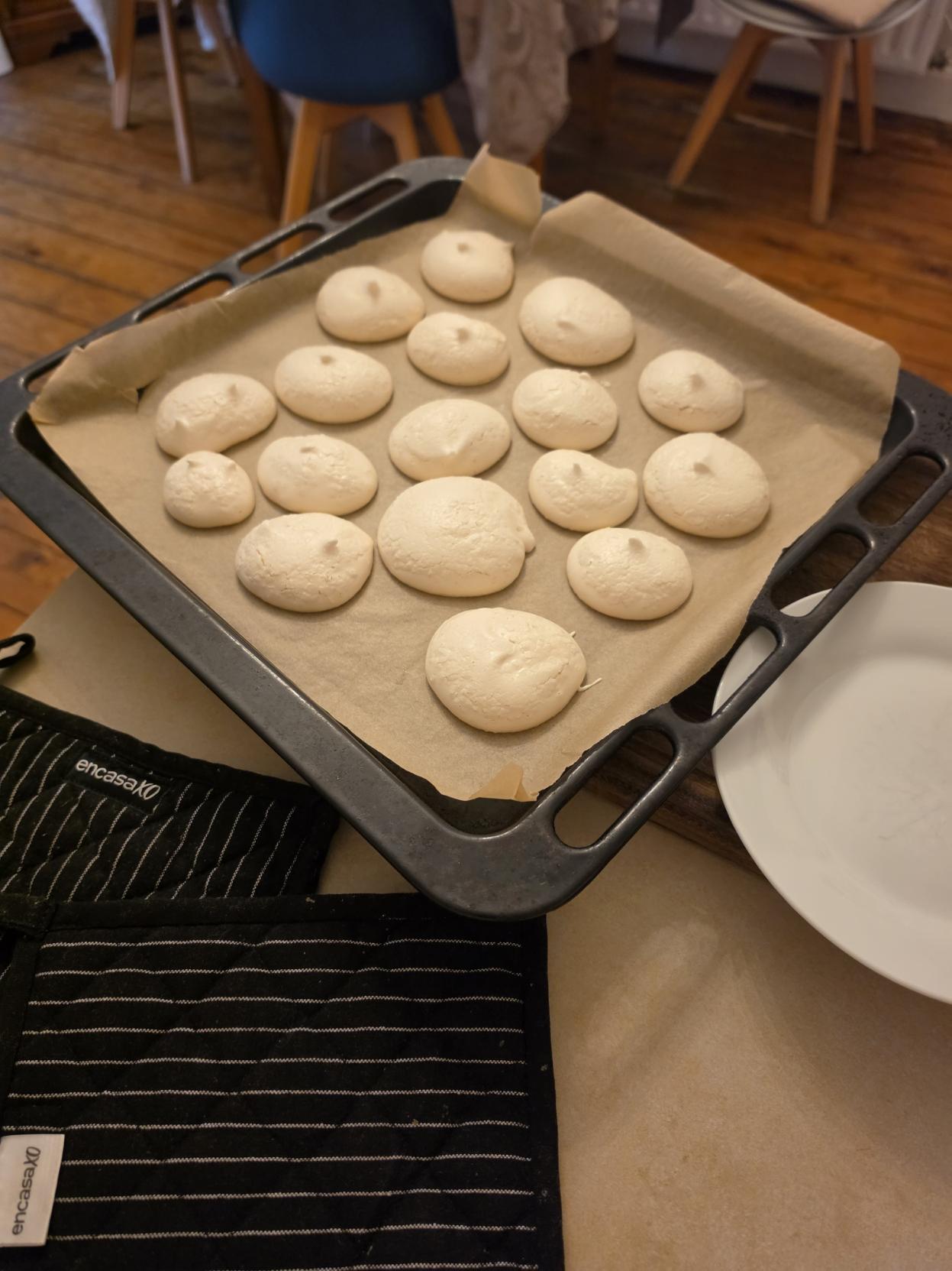A dark metal baking sheet lined with brown parchment paper sits on a kitchen counter, filled with baked, off-white meringue mounds that have a smooth, matte finish with soft, pointed peaks. Next to the tray are black pinstriped oven mitts labeled "encasa" and a empty white ceramic plate. In the background, chairs and wooden floorboards are softly blurred.