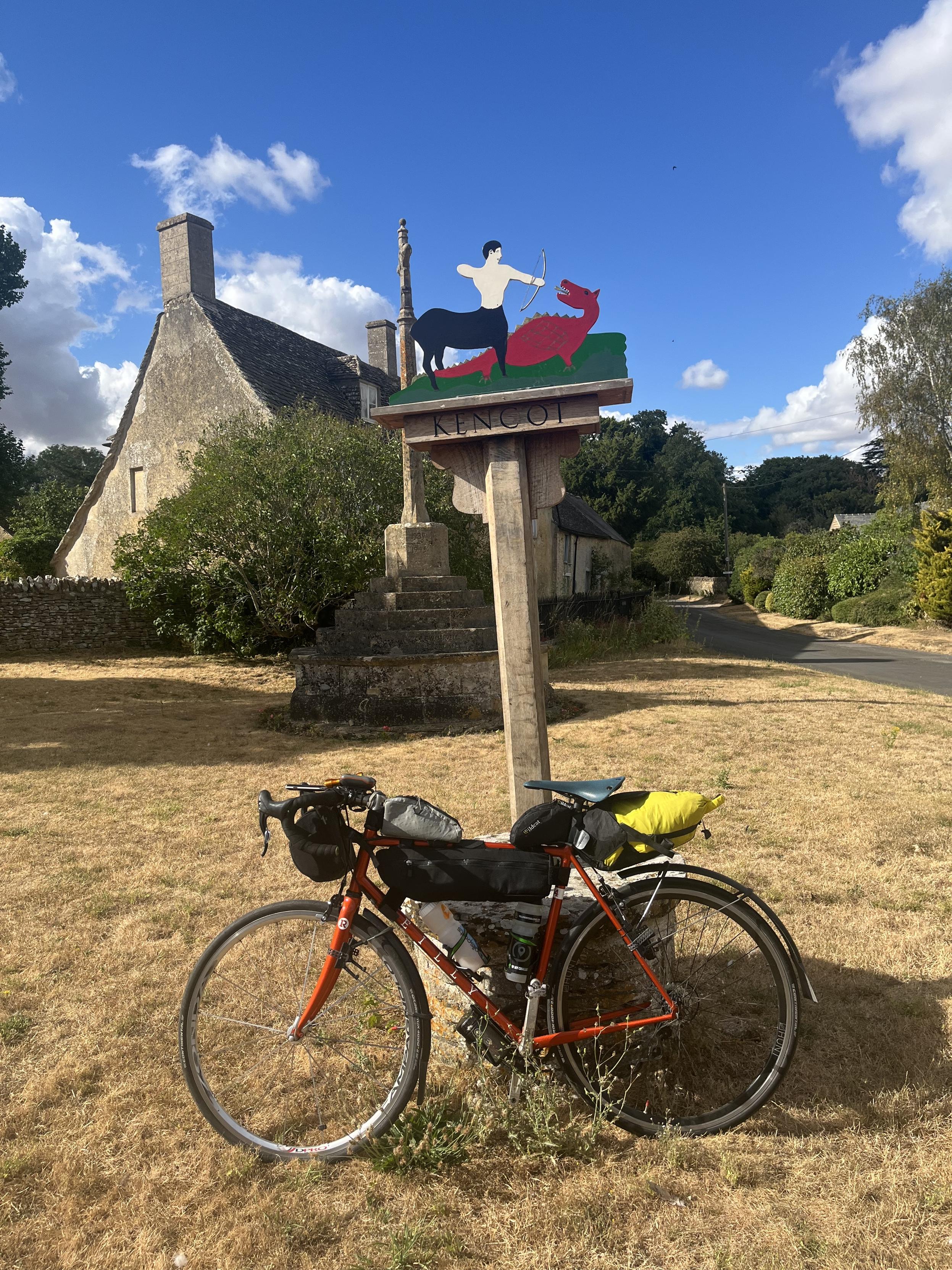 A loaded touring bike leaning on a village sign post with a war memorial and a building beyond