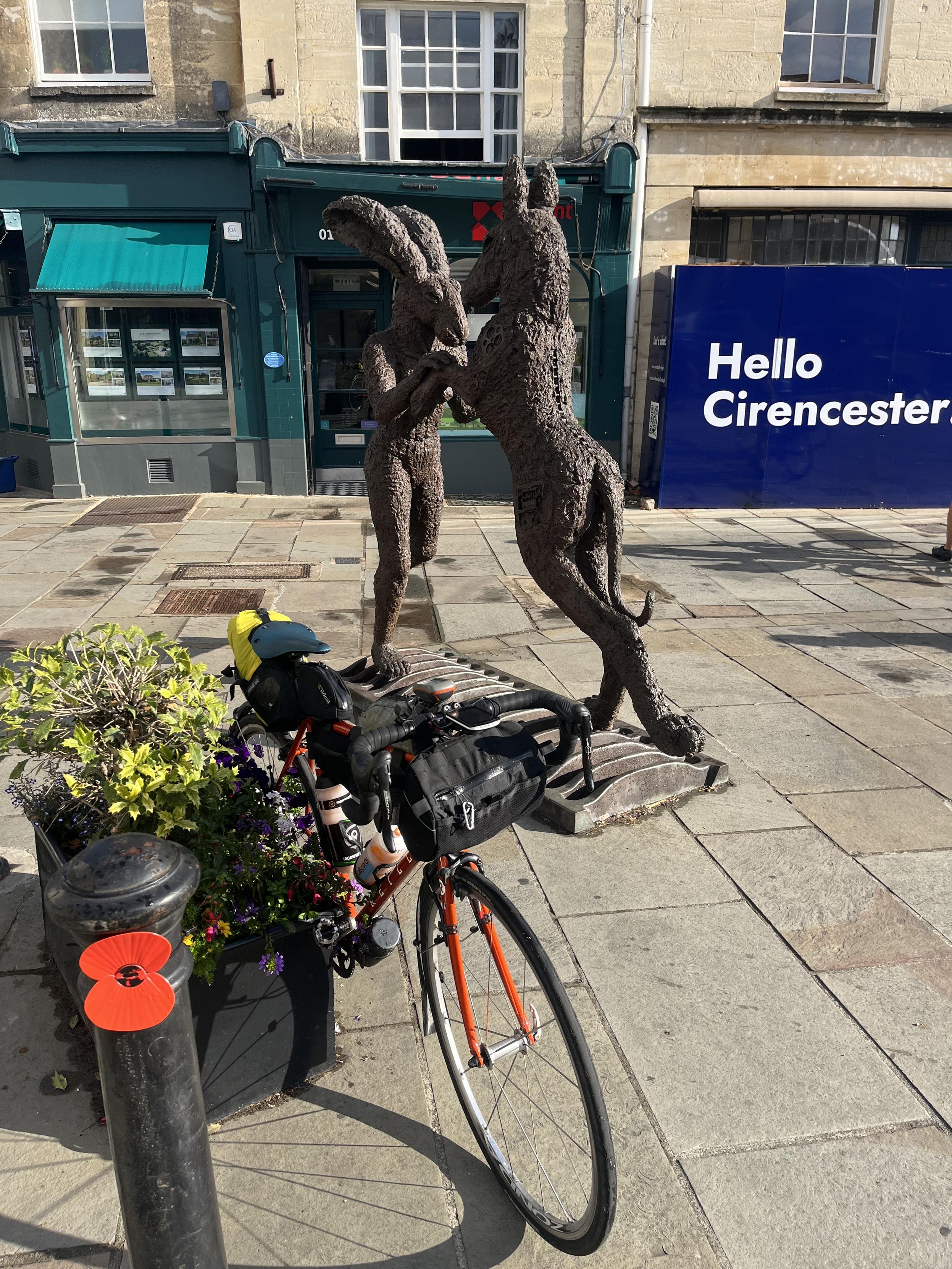A bicycle learnt against a plant display with a hare boxing a dog statue next to a Hello Cirencester sign