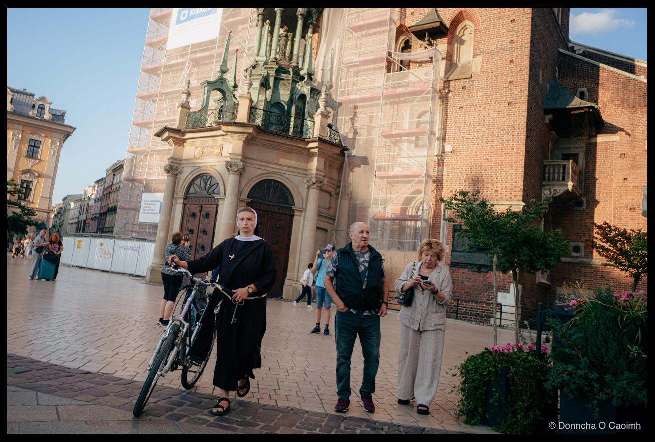 A nun in a black habit and white veil wheels a bicycle across the cobbles of Market Square in Kraków. Behind her rises St Mary's Basilica in red brick and pale stone, its side porch wrapped in scaffolding and green mesh, with a banner reading "Kraków" at the top. A bald man in a dark gilet and a woman in a cream linen suit looking at her phone walk past on the right, and tourists with cameras and a suitcase move through the shade beyond.