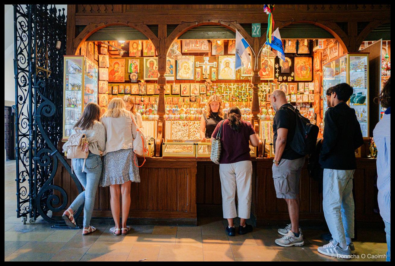 A souvenir stall inside the wooden arcade of the Cloth Hall in Kraków, its back wall covered from counter to arch with gilded religious icons, painted crucifixes and rows of rosaries under warm lights, with a small green plaque numbered 27 above the arch and a flag hanging beside it reading "UKIE". Six shoppers stand at the counter with their backs to the camera, looking down into a glass case of amber and silver jewellery, while a fair-haired stallholder in a black top stands behind it. A black wrought-iron gate with scrolled ironwork fills the left of the frame, and afternoon light falls across the stone floor.