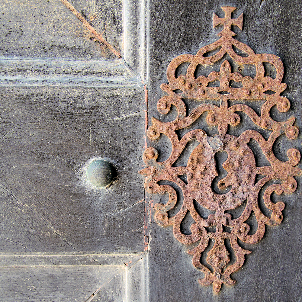 Detail of a church door in Sagres, Portugal showing wood paneling with a small door knob and ornamental decorative ironwork.