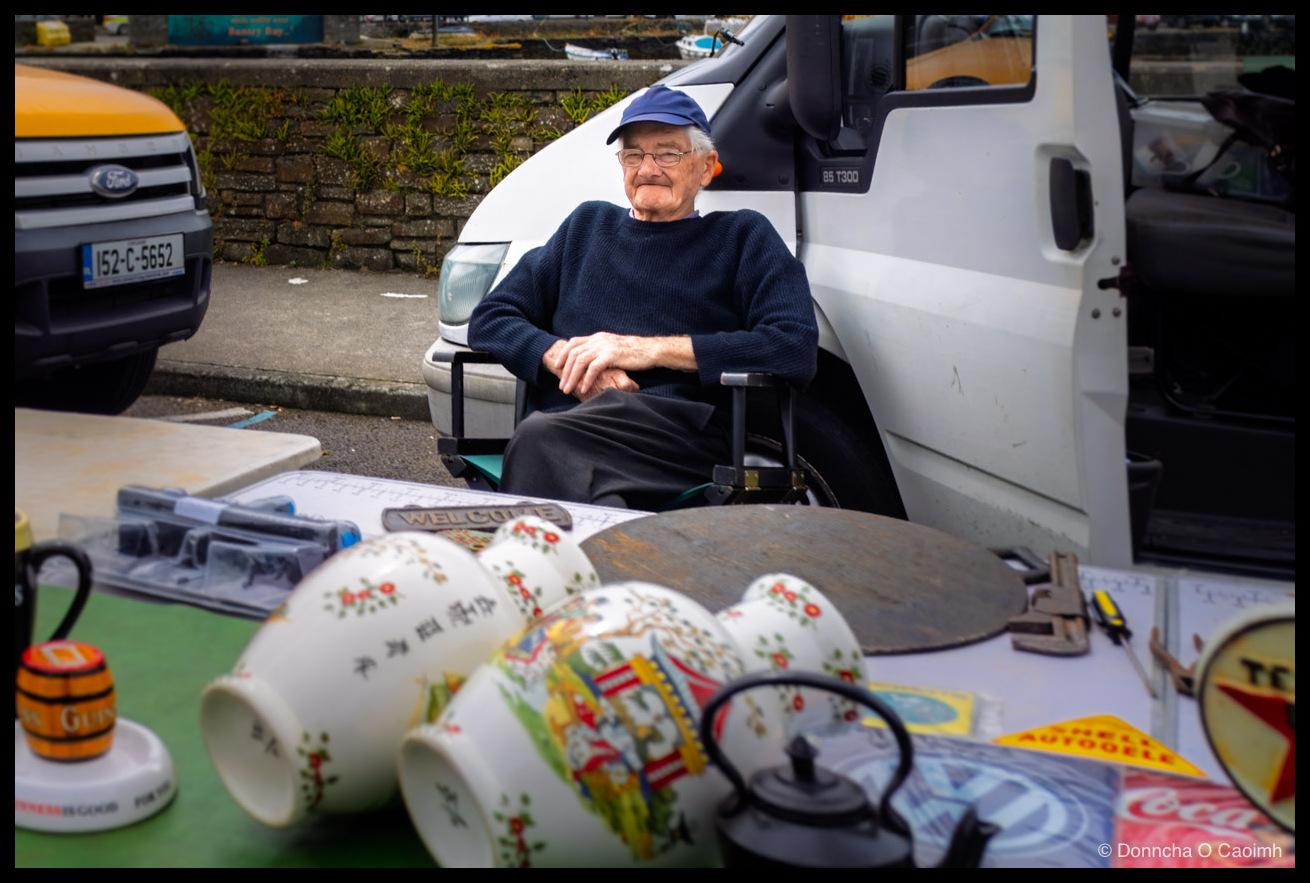An older man in a navy jumper and blue baseball cap sits back in a camping chair with his hands folded, between a white Ford Transit van marked "85 T300" with its side door open and a Ford van registered "152-C-5652", at an outdoor market beside a stone harbour wall in Bantry with boats and a "Bantry Bay" sign behind him, while the foreground table in front of him is crowded with bric-a-brac including a cast metal "WELCOME" plaque, several white patterned jugs, a black cast iron teapot, a Guinness barrel ashtray reading "GUIN" on the barrel and "GOOD FOR YOU" on the base, a large rusted circular metal plate, and enamel advertising signs for "SHELL AUTOOELE", Coca-Cola and Volkswagen.