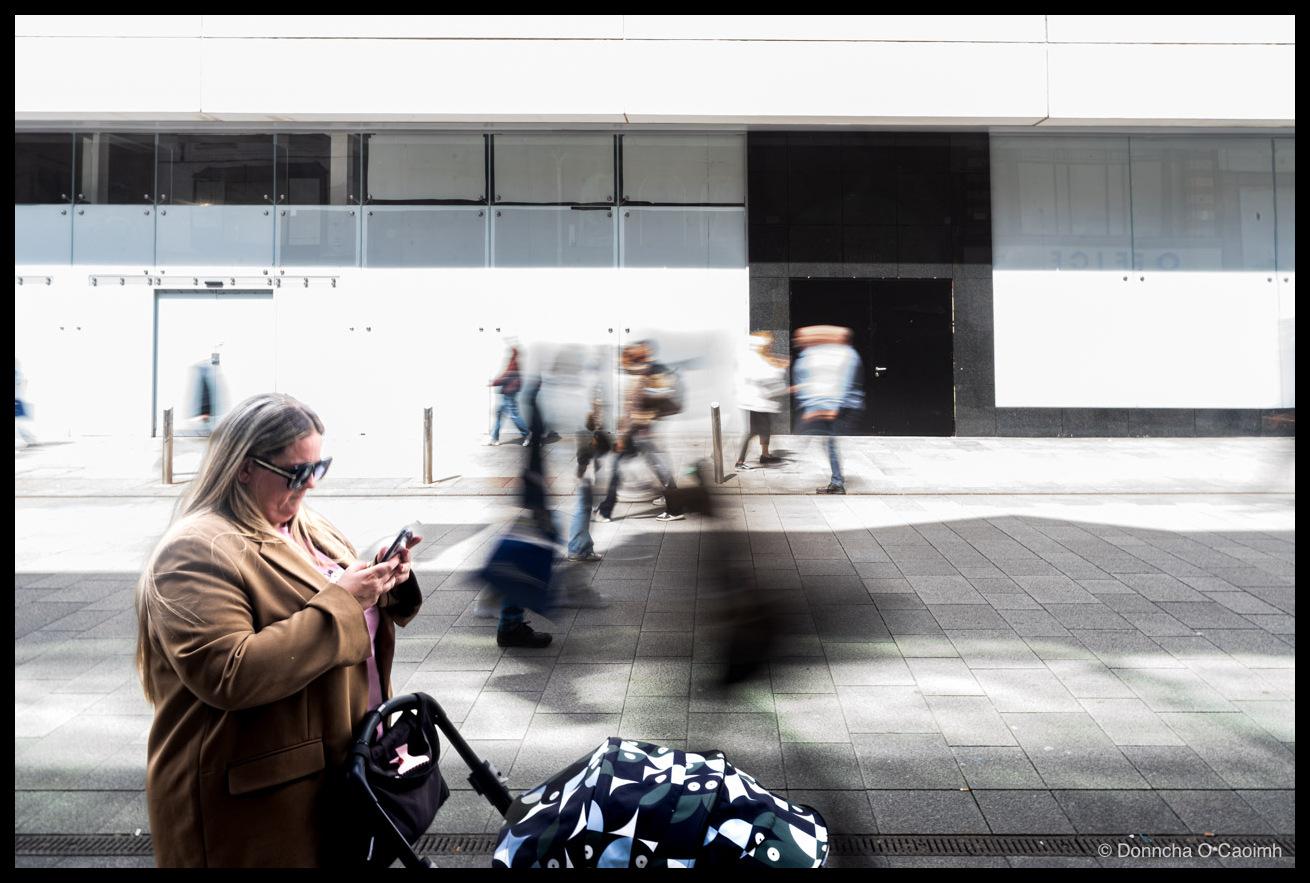 A woman in a camel wool coat and dark sunglasses stands still on a wide granite pavement, looking down at her phone while pushing a black pram with a patterned green, navy and white cover, as blurred pedestrians stream past behind her in front of a boarded-up shopfront of white panels and glass, with faint lettering reading OFFICE on the window to the right.