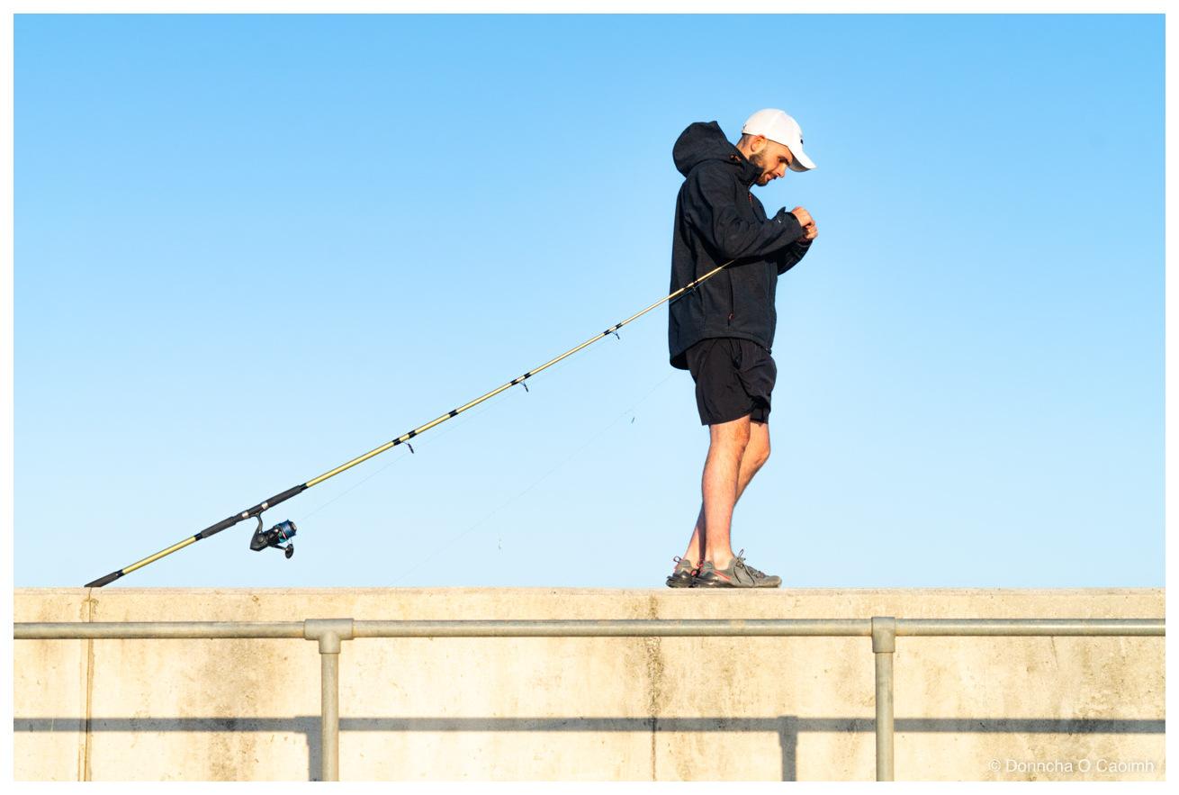 A man in a black hooded jacket, black shorts, trainers and a white baseball cap fixes the line in his fishing rod along the top of a concrete sea wall against a clear blue sky, with a long yellow fishing rod with a spinning reel angled down behind him, and a metal handrail on the wall below.