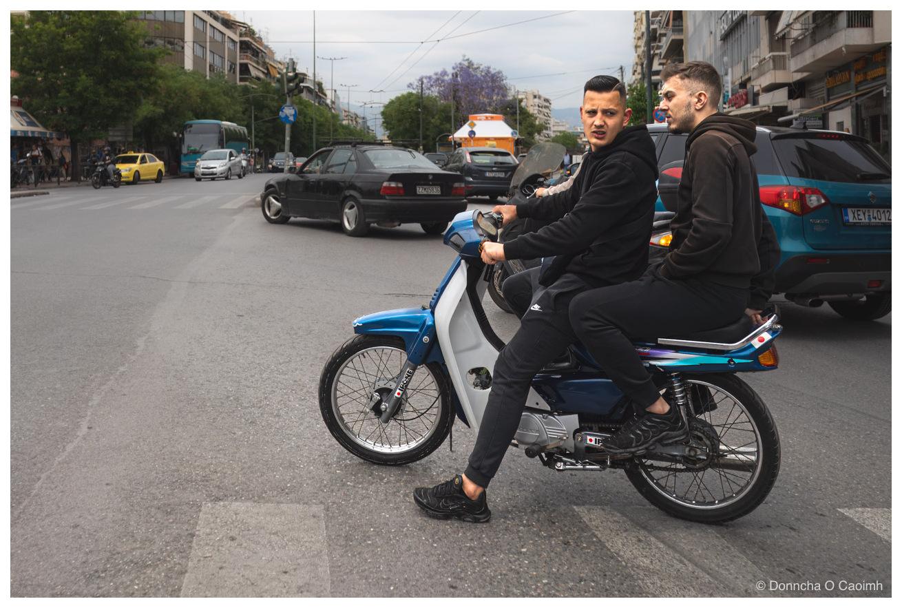 Two young men on a blue and white step-through motorbike waiting at a junction in Athens, the rider in a black hooded top looking back at the camera and his passenger in a brown hoodie looking away down the street, with a black BMW saloon, a yellow taxi, a coach and a teal SUV registered XEY 4012 queuing behind them under overcast sky.