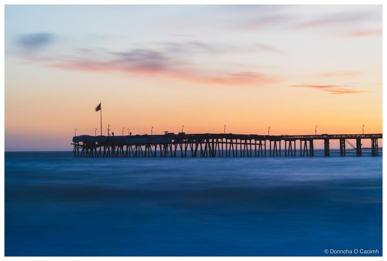 A long wooden pier in silhouette against an orange and blue sunset sky, its trestle legs standing in sea blurred smooth by a long exposure, with a row of lamp posts along the deck and a flag on a pole near the shore end.