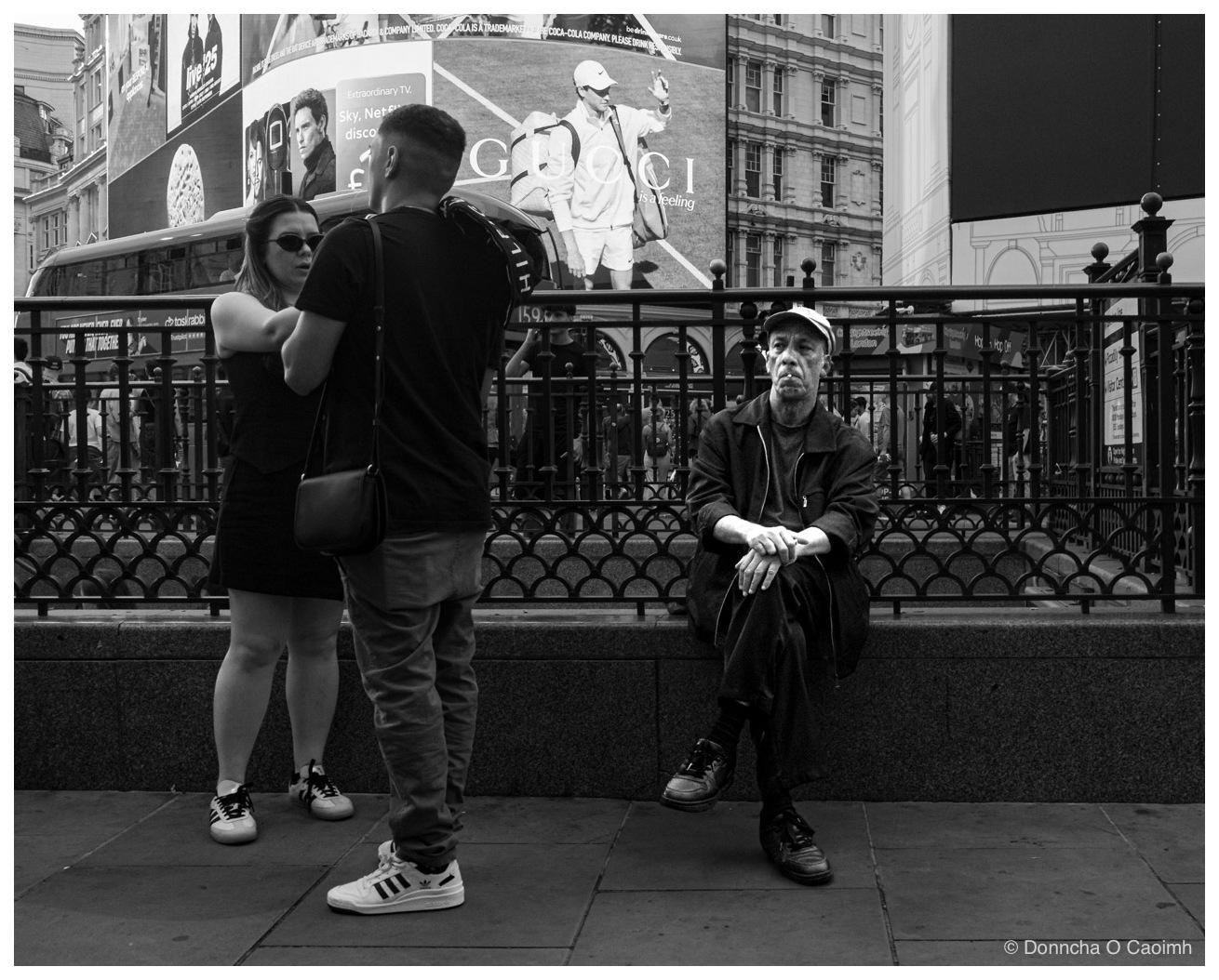 Black and white street scene in Piccadilly Circus, London. A man in a pale flat cap and dark zipped jacket sits on a low granite ledge in front of ornate iron railings, hands clasped over his knees, looking towards the camera. In the foreground a woman in a black dress and sunglasses, with a small shoulder bag, stands talking to a young man in a black T-shirt and jeans with a holdall on his shoulder and white Adidas trainers. Large advertising screens fill the background, showing "live 125", "Extraordinary TV. Sky. Netflix", a tennis player carrying a bag beside the word "GUCCI" and "is a feeling", and a curved Coca-Cola panel reading "COCA-COLA IS A TRADEMARK OF THE COCA-COLA COMPANY. PLEASE DRINK RESPONSIBLY. be.drinkaware.co.uk". Smaller signs behind the railings read "taskrabbit", "Trustpilot" and "Hop On Hop Off".