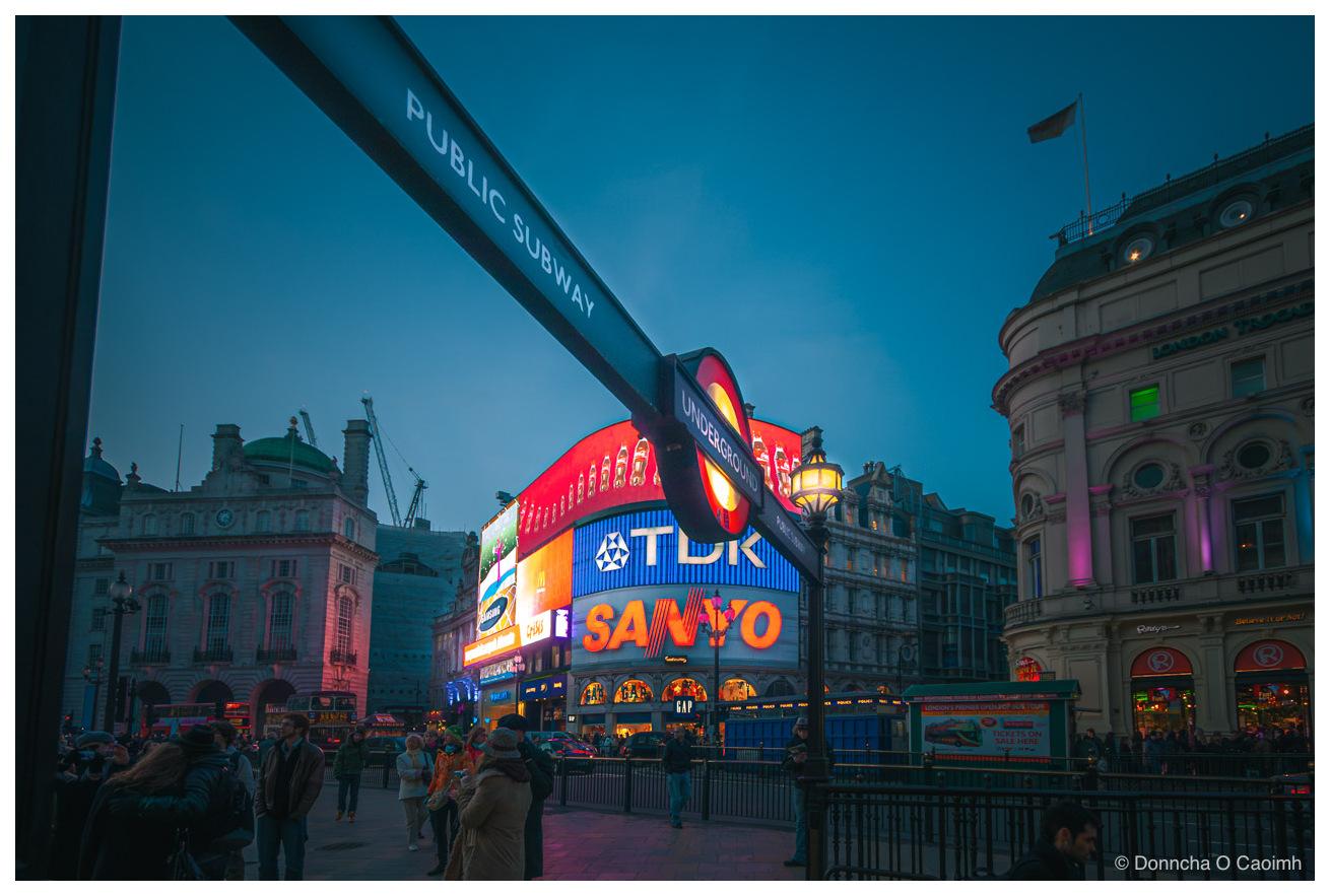 Piccadilly Circus at dusk, with a black metal sign reading "PUBLIC SUBWAY" angled across the top of the frame and the red, white and blue London Underground roundel below it, the illuminated Coca-Cola curved advertising screens behind showing "TDK", "SANYO" and "SAMSUNG", a GAP store at street level, the London Trocadero and Ripley's Believe It or Not! building on the right with a hoarding reading "LONDON'S PREMIER OPEN TOP TOUR" and "TICKETS ON SALE HERE", and people standing in coats on the pavement in the foreground.