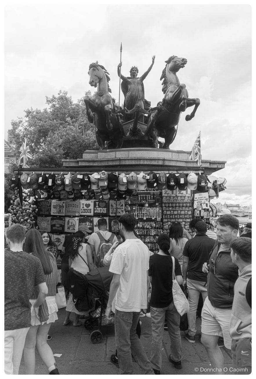 Black and white street scene at the Boadicea and Her Daughters statue in London, where a souvenir stall built against the plinth sells baseball caps, keyrings and fridge magnets under signs reading "LONDON ENGLAND", "I ♥ LONDON" and "WE ACCEPT ALL MAJOR CREDIT CARDS VISA", while a crowd of tourists browses below the rearing bronze horses and the spear-raised figure of Boudicca.