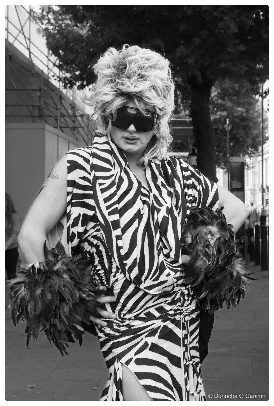 A woman in a zebra-print dress and matching wrap stands with her hands on her hips on a London pavement, wearing a tall blonde bouffant wig, oversized wraparound black sunglasses and black feather cuffs at her wrists, with scaffolding and a plane tree behind her, in black and white.