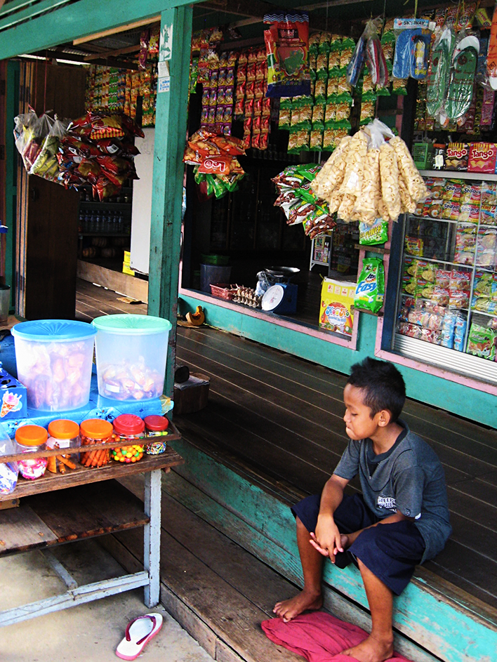 A young boy sits on the steps of a small village shop with a colorful display of food items in the window and hanging on the outside in front of the window and at the entrance. The boy may or may not be minding a display table sitting in front of the shop with an assortment of jars and translucent plastic buckets containing a variety of sweets.