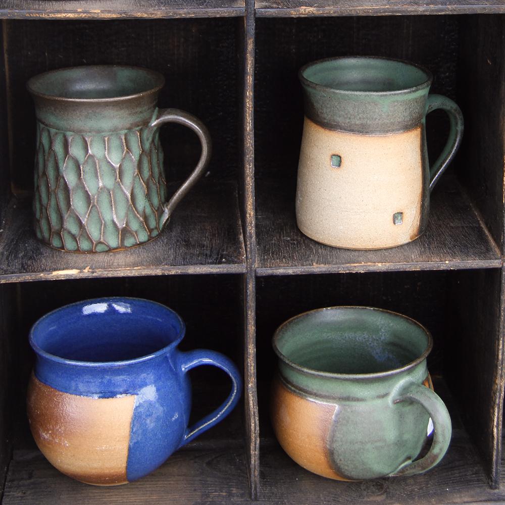 Four stoneware mugs in a rough wooden display cabinet.