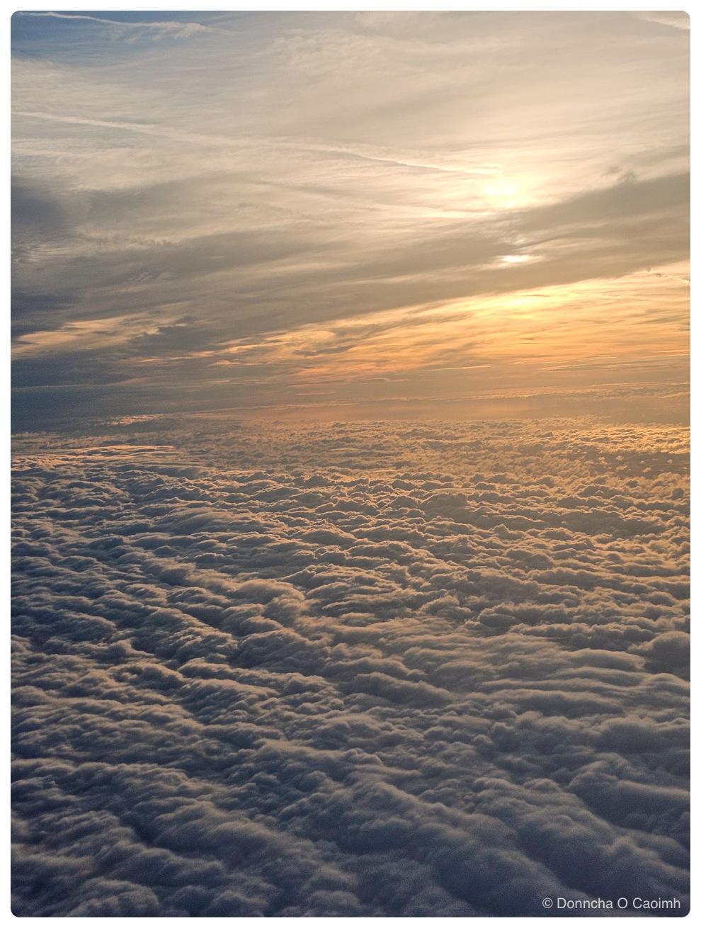 Aerial view from an aircraft window at sunrise, looking out across an unbroken layer of low cloud that is rippled into long ridges running towards the horizon, lit gold and pink by a sun sitting just above the cloud tops behind streaks of higher cloud.