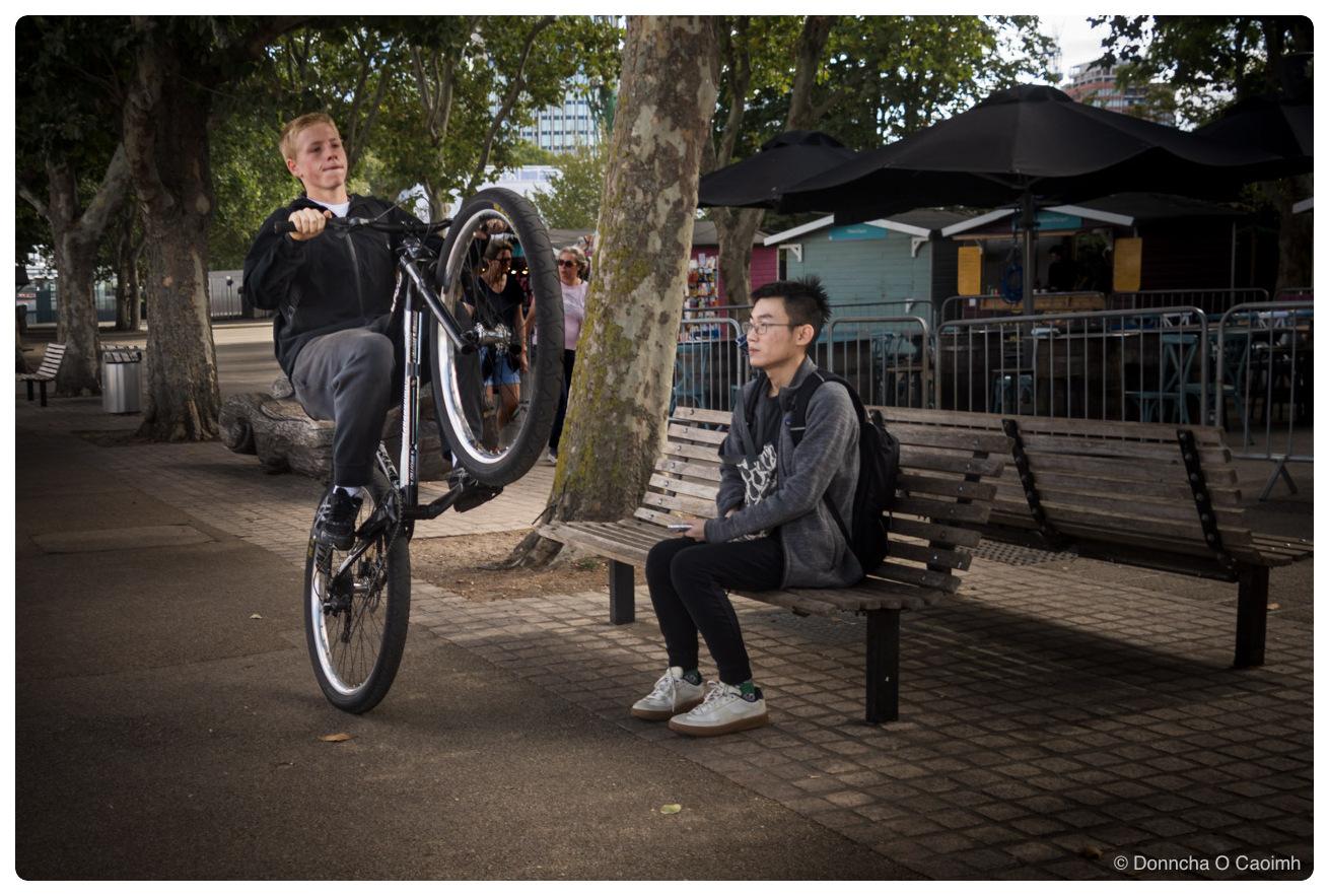 A teenage boy in a black jacket and grey tracksuit bottoms rides a bicycle in a wheelie along a paved riverside path, front wheel raised level with his chest, while a young man in a grey hooded top and black jeans sits on a wooden bench a few feet away looking off to one side, with London plane trees, black café parasols, metal crowd barriers and a pale blue kiosk behind them.