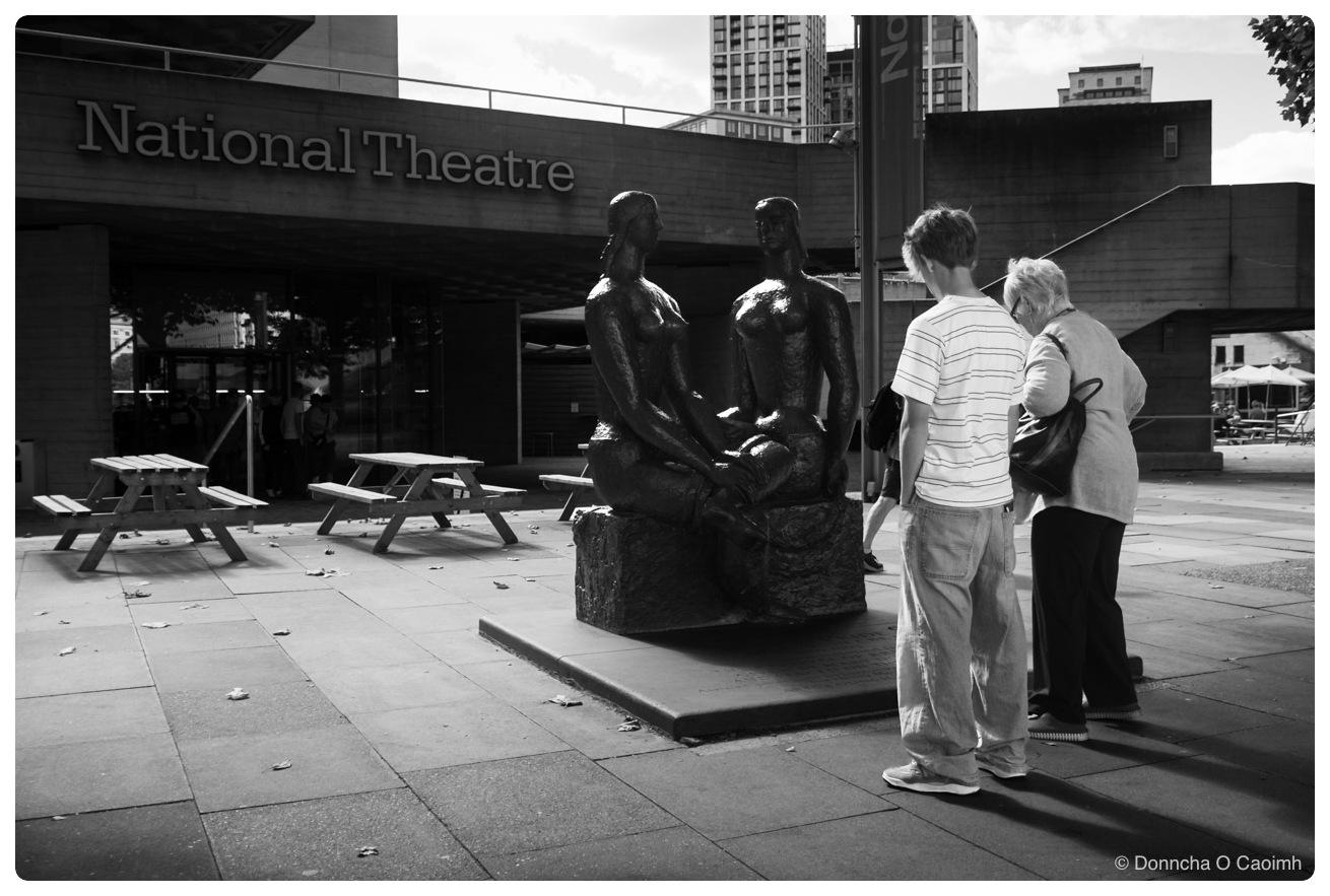Black and white shot of two people standing with their backs to the camera, looking down at a bronze sculpture of two seated female figures on a rough stone base, outside a concrete building with large lettering reading "National Theatre"; empty wooden picnic tables sit on the paving to the left, and tower blocks rise behind the building.