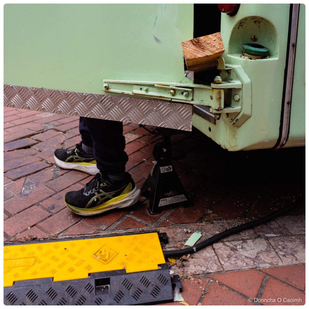 Close crop of the rear corner of a mint green food truck jacked up on a black axle stand, with a chequer plate step, a block of untreated timber wedged into the open door frame, and a person's lower legs in black trousers and black ASICS trainers with yellow soles standing on red brick paving, a yellow and black cable ramp running across the foreground.