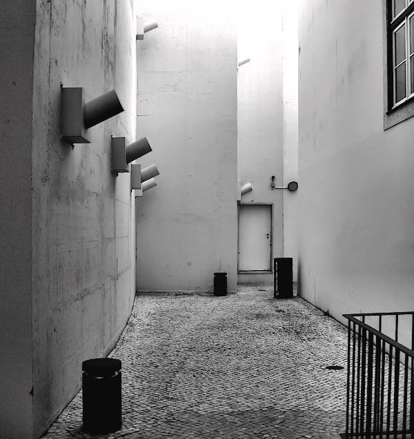 Black and white photo of a cobblestoned long, narrow urban yard with high white walls, a single window on the right, some railings and waste bins, ventilation shafts on the left, and a door at the far end.