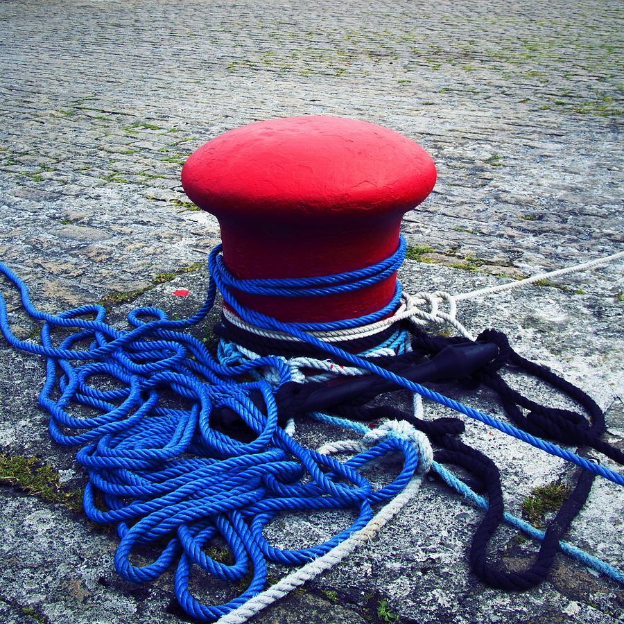 A freshly painted, bright red bollard in a harbour is surrounded by somewhat tangled bright blue, black and white ropes.