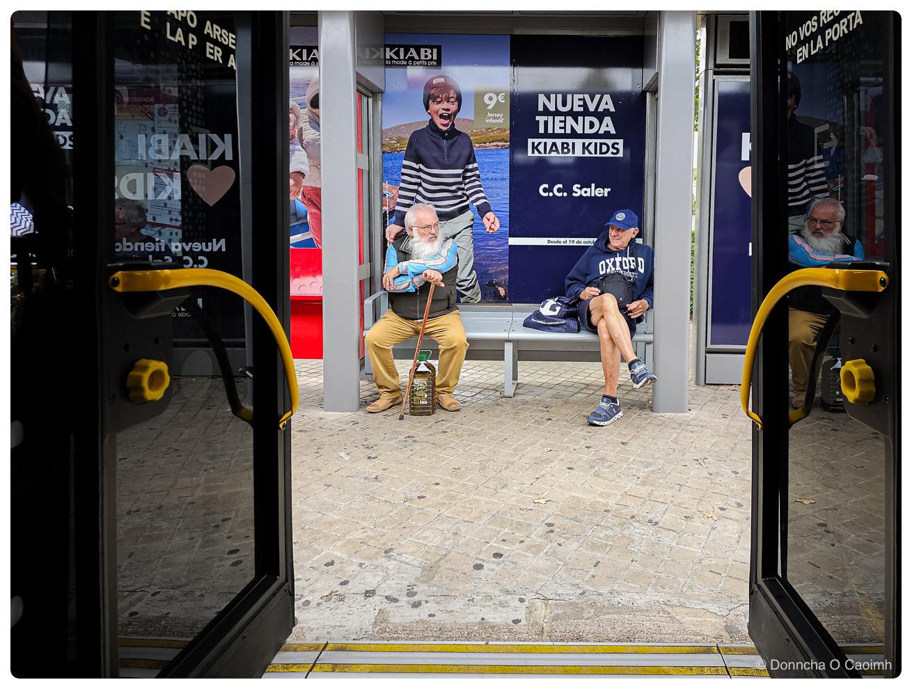Two older men sitting on a bus shelter bench, seen from inside a bus through its open double doors, one in a white beard, glasses, olive gilet and tan trousers with a walking stick and a five litre bottle of olive oil at his feet, the other in a navy cap, an OXFORD sweatshirt and shorts with his legs crossed, behind them a Kiabi advert reading KIABI, la mode à petits prix, 9€ Jersey infantil, NUEVA TIENDA KIABI KIDS, C.C. Saler and Desde el 19 de octubre, with Valencian lettering on the door glass including NO VOS REC and EN LA PORTA.