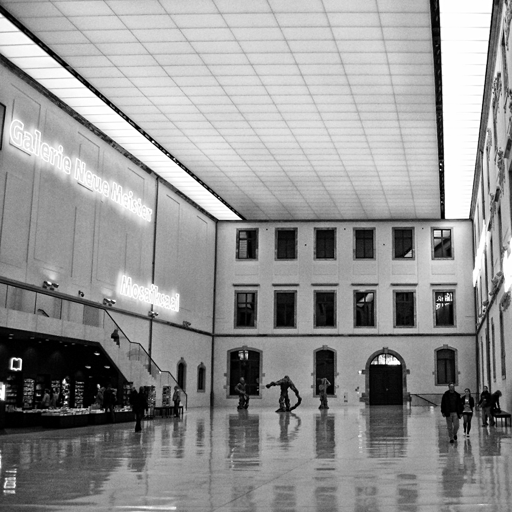 Black and white photograph of the central atrium of the Albertinum, an art museum in Dresden, Germany. The atrium is a very large rectangular space, painted solely white and incorporates two formerly external walls with windows and doorways. The polished floor mirrors those structures as well as visitors and a scuplture. Brightly lit signage denotes the stariwayed entrance to the New Masters Gallery.