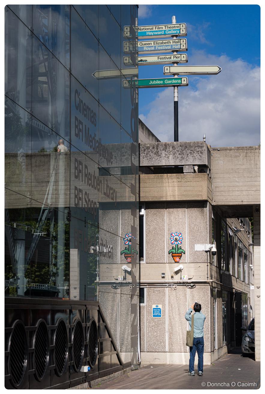 A man in a light blue shirt and jeans, with a green bag over his shoulder, photographs a pixel-art tile mosaic of flowers in an orange pot with red, white and blue petals on a concrete wall on London's South Bank, beside real CCTV cameras mounted on the wall. To the left, a glass wall reflects the surroundings and reads "Cinemas", "BFI Mediatheque", "BFI Reuben Library", "BFI Shop" and "BFI bar & kitchen". Above, a signpost against a blue sky with white clouds points to "National Film Theatre", "Hayward Gallery", "Queen Elizabeth Hall", "Royal Festival Hall", "Festival Pier" and "Jubilee Gardens", and a blue sign on the wall reads "Keep clear Exit from emergency escape route".