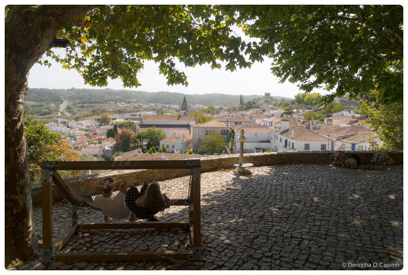 A couple lie side by side in a rope net hammock strung inside a wooden frame on a cobbled terrace on the town walls of Óbidos, shaded by a large plane tree, with whitewashed houses, terracotta roofs and a church spire below and wooded hills in the distance.