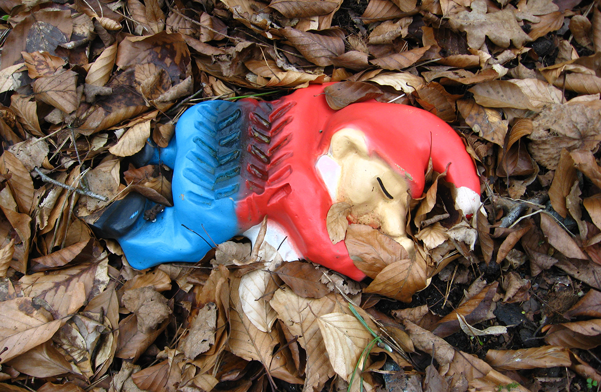 A satirical take on kitschy garden gnomes: here a garden gnome lies squished dead in leaf litter with tire tracks running over his body.