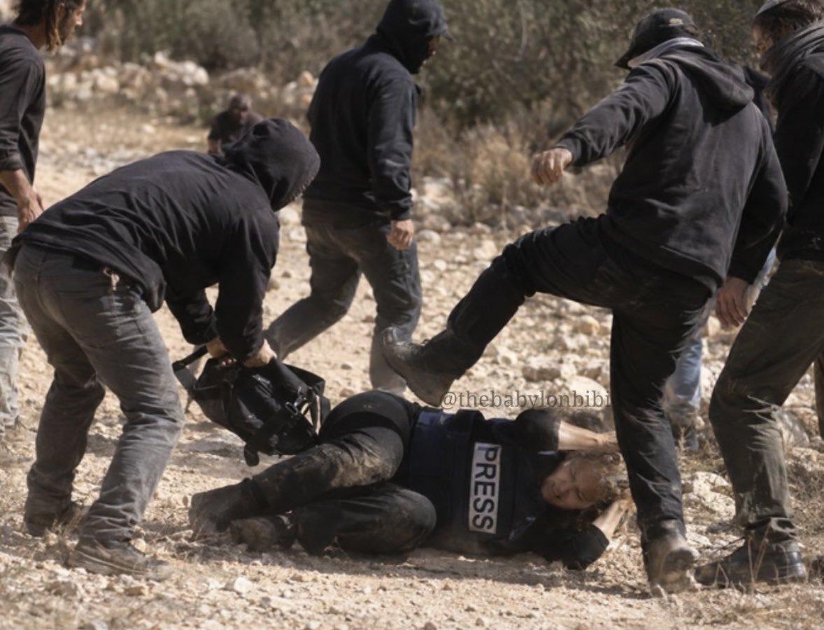A group of people in dark clothing are gathered around a person lying on rocky, dirt ground. The person on the ground is wearing a dark vest with the word "PRESS" printed in white. One person in the foreground leans over the person on the ground near black equipment. Another person has a leg raised toward the person on the ground with a clenched fist. The background consists of a dry, hilly landscape with sparse, leafless vegetation.