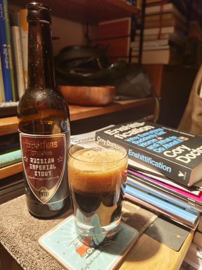 A small glass with dark brown beer in the lower middle, left side behind the bottle of beer one to the right a stack of magazines with a book upon. The book is titled "Enshitification". All on a small table in front of bookshelves.