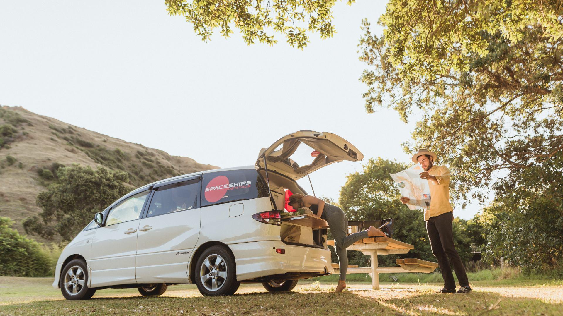 Spaceships campervan is parked at a campsite - green forest/hilly area. One Space Traveller is getting something from the boot while the other one is looking at a map. It is a sunny day.
