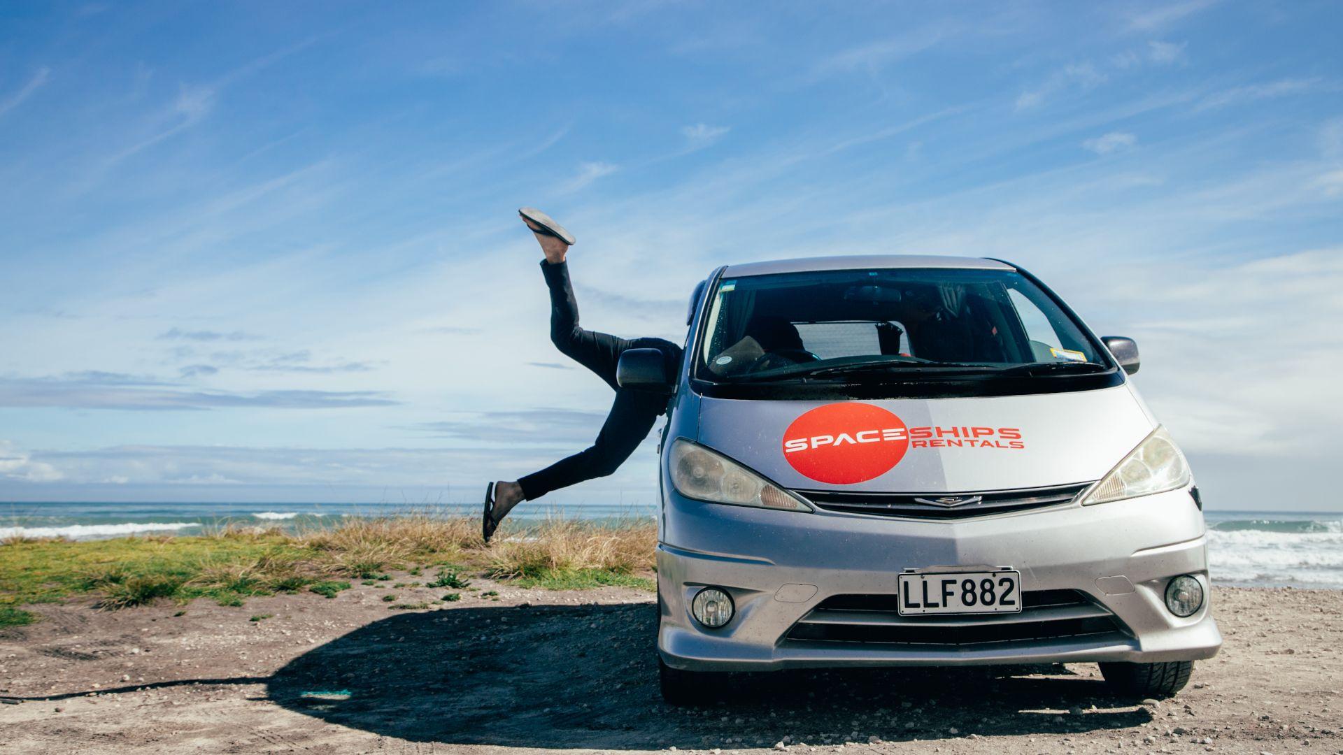 Spaceships campervan is parked facing forward. Space Traveller is jumping through the open side window as they don't want to miss out on an amazing road trip. They are ready to go. Beachy area with ocean in the background on a sunny day.