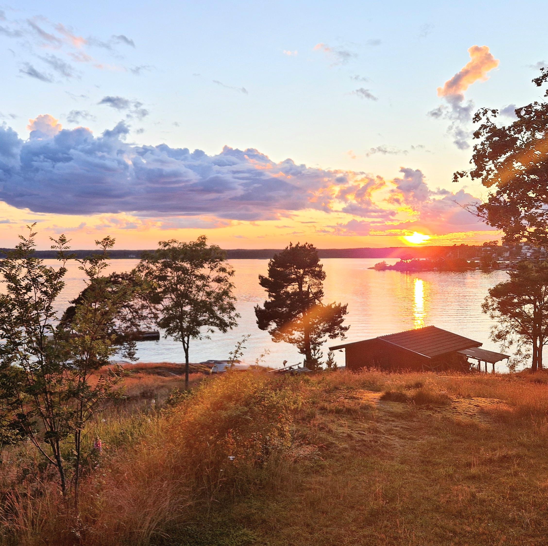 Ljus himmel med några mörka moln. En sol på väg att gå ned bakom trädtopparna. Solstrimman speglar sig i sjön. I förgrunden torrt gräs, några träd och en sjöbod.