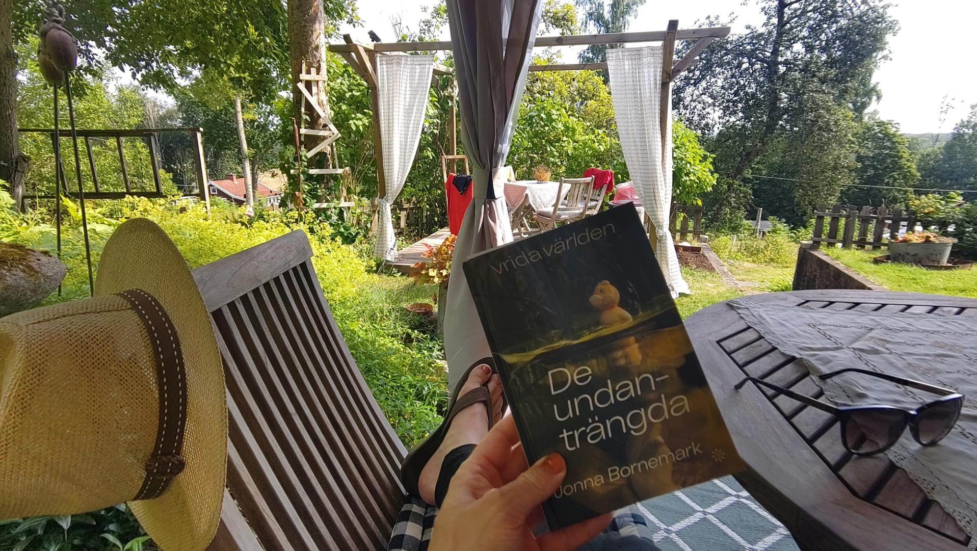En bild på en sommaridyll, med trästolar, grönska och sommarhatt. En hand som håller i boken "de undanträngda" av Jonna Bornemark