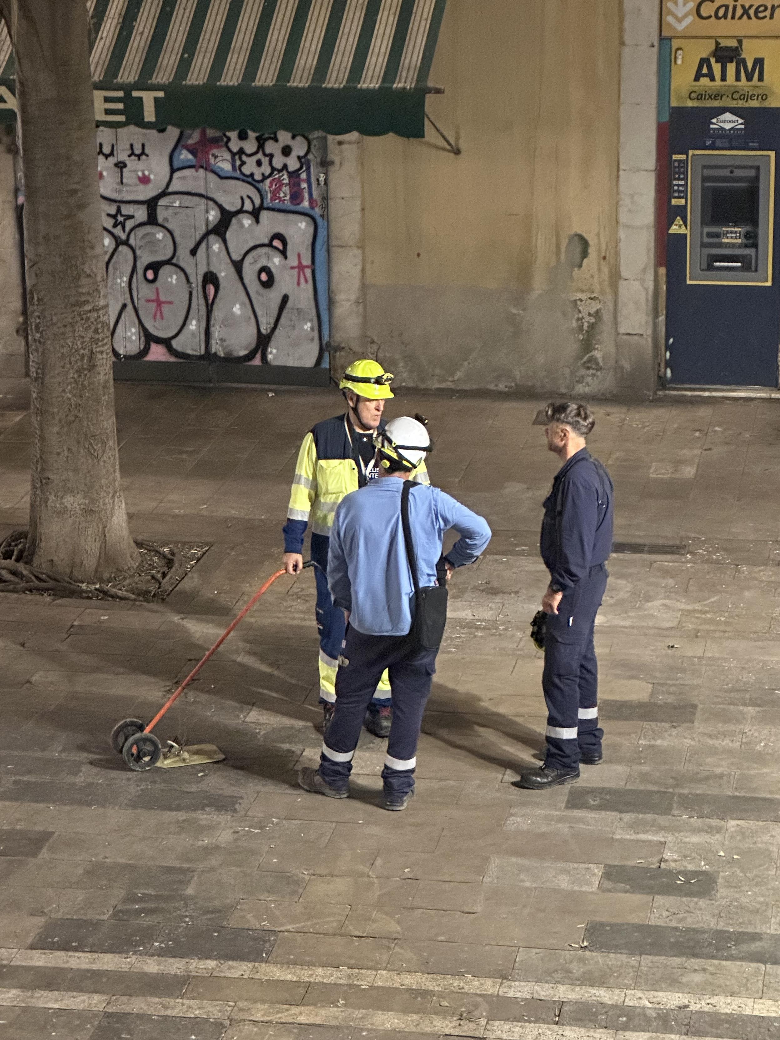 Tre män står och snackar på ett torg. Man ser ett verktyg med hjul och orange handtag som en av männen lutar sig mot.
