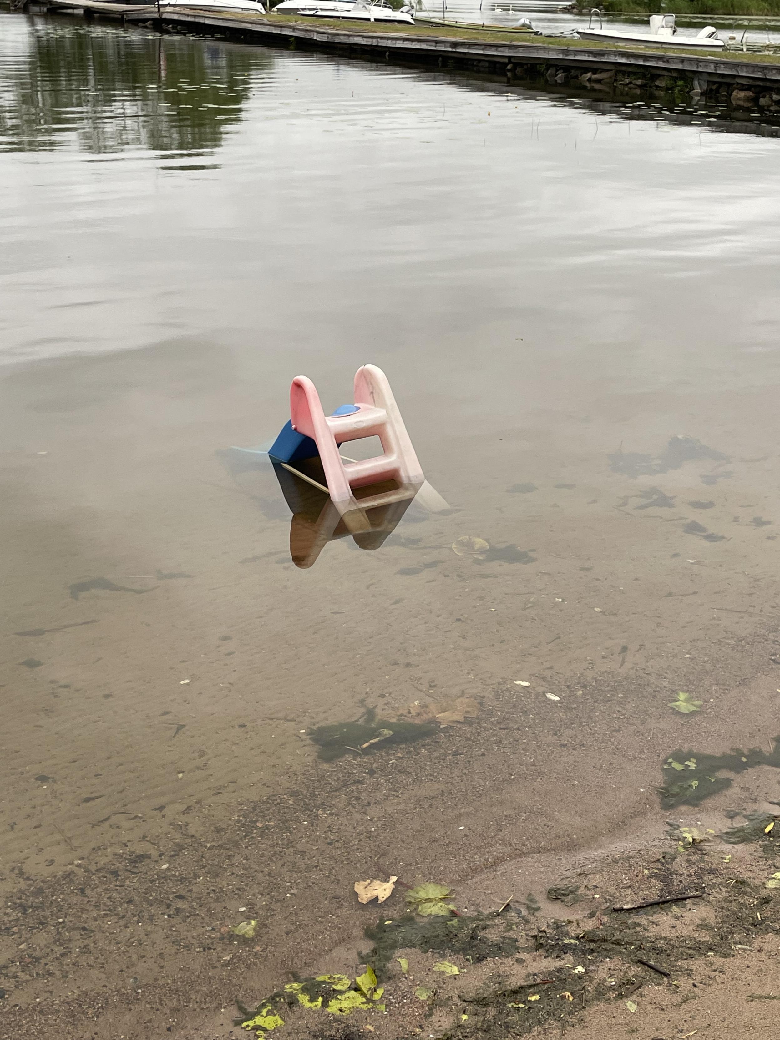 En väldigt liten, solblekt rutschkana i plast. Den är placerad någon meter ut från strandkanten och ser övergiven ut. Vattnet är klart så man ser den bruna botten.