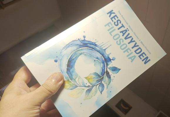 Kirja kädessä. Kannessa tyylitelty vesivärimaalattu pyörivä maapallo, joka muodostuu  toisella puolella loiskuvasta vedestä ja toisella puolella kaarelle taipuvan varvun lehdistä