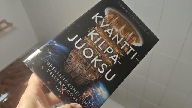 Kirja kädessä. Kannessa otsikon alla kattokruunun kaltainen kvanttitietokone.
